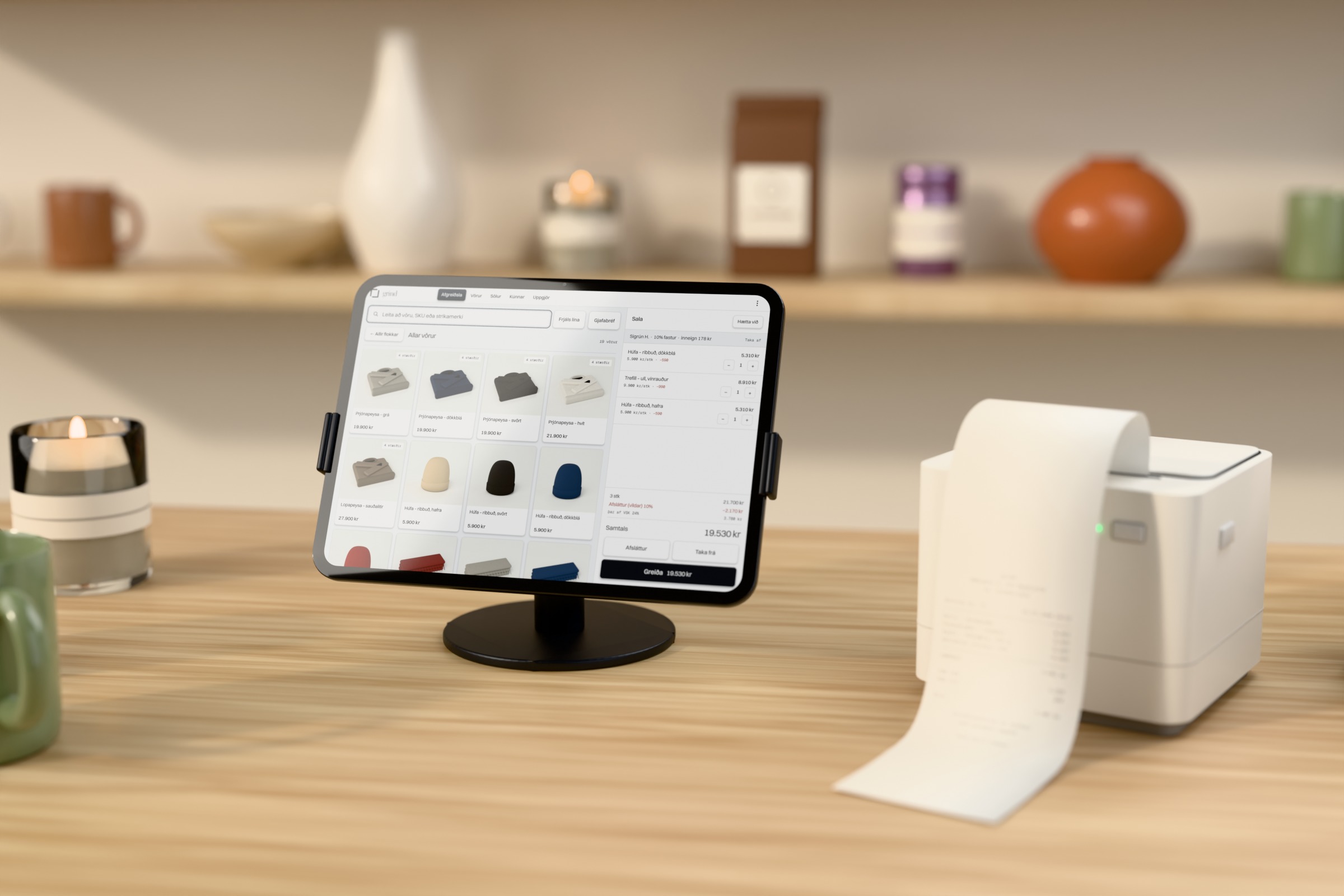 iPad á dökkum armstandi á eikarborði í búð, með grind-kassann á skjánum og hillu með vörum fyrir aftan, hvítur kvittanaprentari við hliðina með kvittun sem rennur út á borðið, og bolli og ilmkerti í forgrunni.